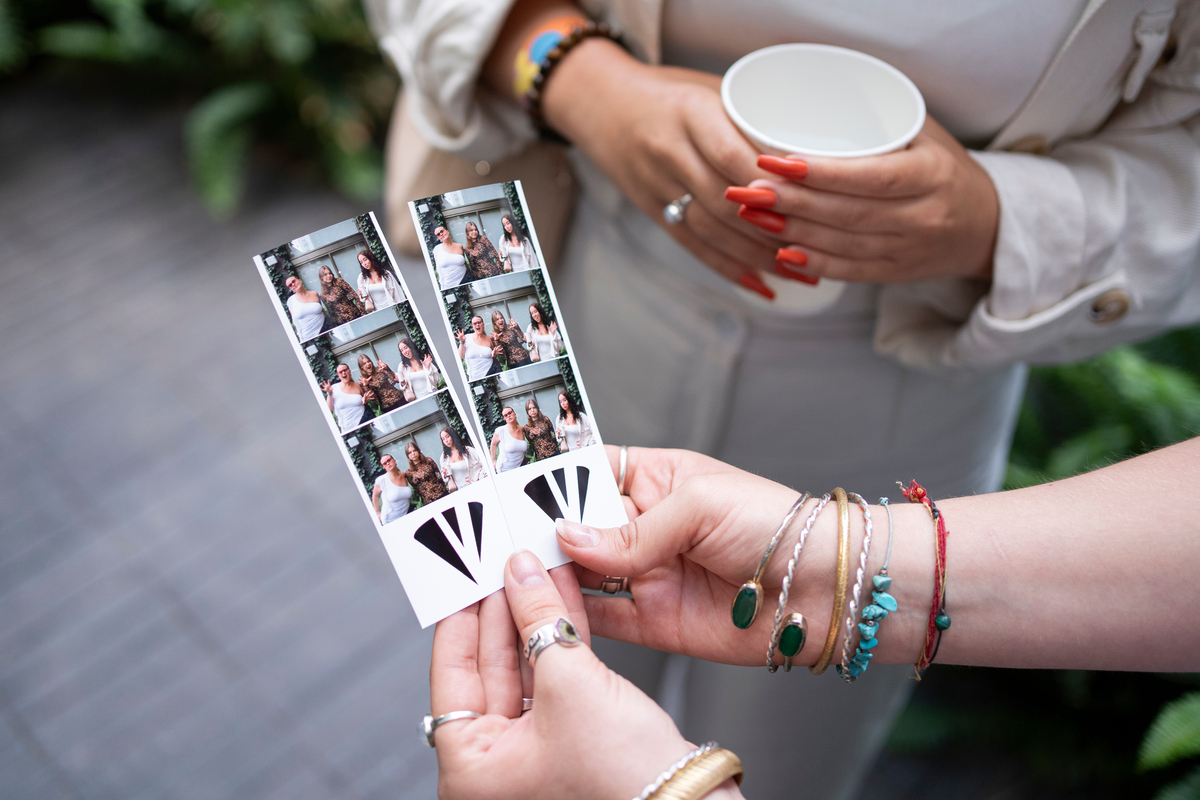 Two people, dressed in light-colored outfits, are holding photo booth strips of group photos. One is holding a cup, and both have colorful bracelets. They are standing on a tiled surface with green foliage in the background.